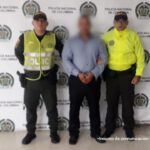 En la imagen aparece el procesado con camisa blanca azul y pantalón negro custodiado por dos agentes de la Policía Nacional delante de un pendón de esa institución.