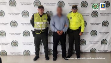 En la imagen aparece el procesado con camisa blanca azul y pantalón negro custodiado por dos agentes de la Policía Nacional delante de un pendón de esa institución.