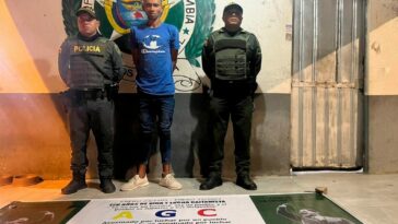 En la imagen se observa a un hombre de camiseta azul con jean y tenis blanco, custodiado por dos agentes de la Policía Nacional. Delante suyo se observa el pendón de las Autodefensas Gaitanistas de Colombia que supuestamente estaba colgando en una calle.