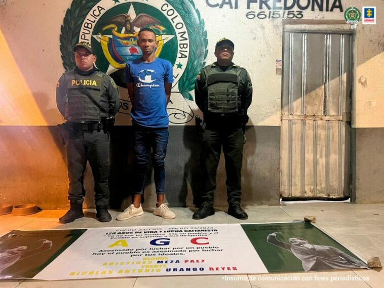 En la imagen se observa a un hombre de camiseta azul con jean y tenis blanco, custodiado por dos agentes de la Policía Nacional. Delante suyo se observa el pendón de las Autodefensas Gaitanistas de Colombia que supuestamente estaba colgando en una calle.