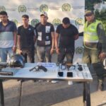 En la imagen se observa a cuatro hombres custodiados por dos agentes de la Policía Nacional, detrás de una mesa donde se identifican un arma de fuego, municiones y celulares.
