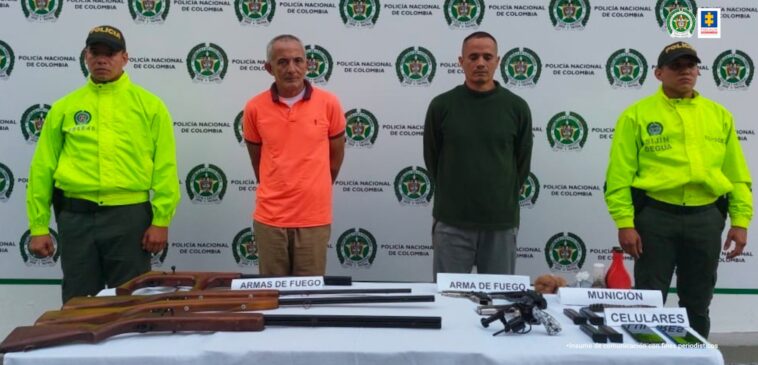 En la imagen se ven dos personas detenidas bajo custodia de la Policía Nacional. Detrás suyo un backing de la institución y delante de ellos una mesa con armas y otros elementos incautados.
