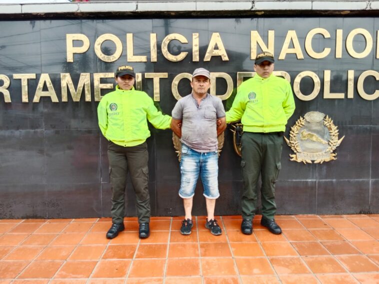 En la fotografía se ven tres personas una de ellas el capturado, acompañados de dos uniformados de la Policía Nacional. En la parte de atrás se observa el aviso de entrada de la Policía.