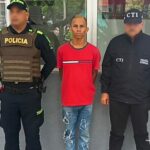 En la fotografía aparece un hombre de pie vestido de camiseta roja y jean azul,  custodiado por un uniformado del CTI de la Fiscalía y uno de la Policía Nacional.