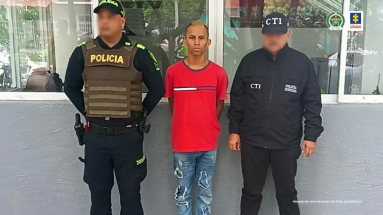 En la fotografía aparece un hombre de pie vestido de camiseta roja y jean azul,  custodiado por un uniformado del CTI de la Fiscalía y uno de la Policía Nacional.