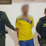 El capturado viste una camiseta de la selección Colombia, está esposado con sus manos a la espalda y está custodiado por dos uniformados de la policía nacional.