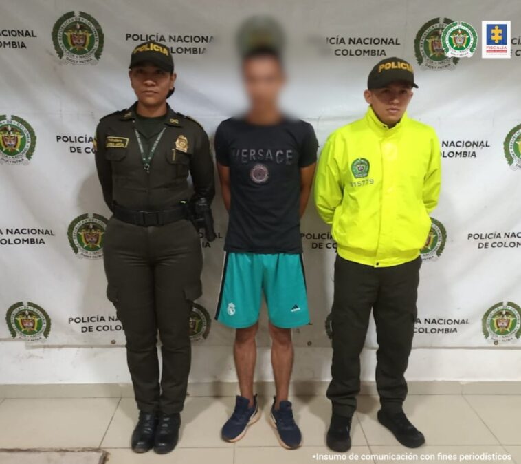 El capturado está en medio de dos uniformados de la policía nacional, tiene una pantaloneta verde y una camiseta negra y sus manos esposadas a a la espalda.