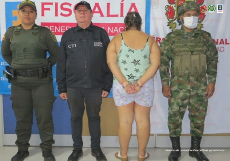 capturada de espaldas esposada custodiada por militar, investigados del CTI de la Fiscalía y un policía. Detrás banner de la Fiscalía y del Ejército.