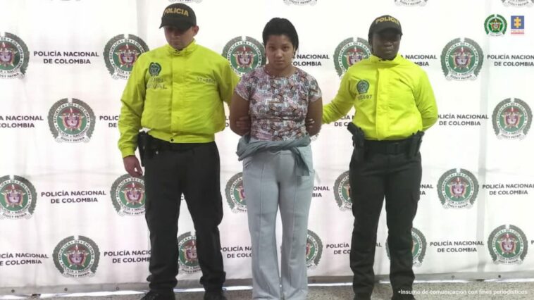 En la fotografía está la procesada con una camiseta, una sudadera gris y un saco amarrado a la cintura con dos servidores de la Policía Nacional que usan chaqueta verde fluorescente y al fondo hay un pendón de la Policía con sus logos en mosaico.