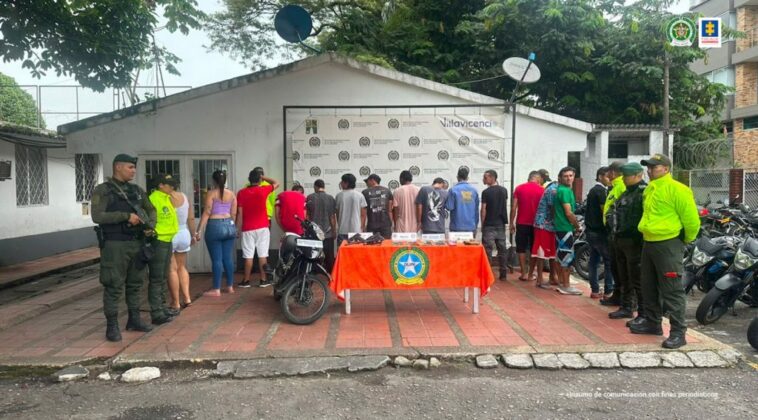 capturados de espaldas custodiados por policias, delante de ellos una mesa con material incautado y una moto. Detrás banner de la Policía Nacional.