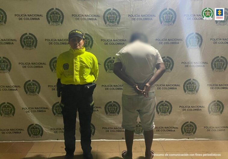 En la fotografía hay un hombre de pie, de espalda, vestido de bermuda y camiseta beiges, acompañado de un uniformado de la Policía Nacional.