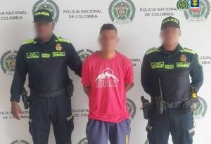 En la imagen aparece un hombre vestido con camiseta roja y pantalón corto azul, está de pie, con el rostro difuminado, los brazos esposados ​​a la espalda, acompañado de dos miembros uniformados de la Policía Nacional.